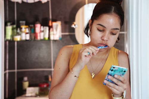 Mixed race young woman, brushing teeth and checking phone outside bathroom