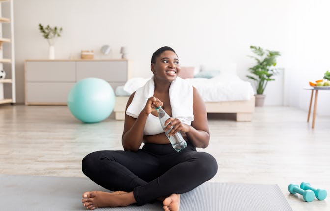 Plump black lady in sportswear sitting on sports mat with bottle of clear water at home, full length...